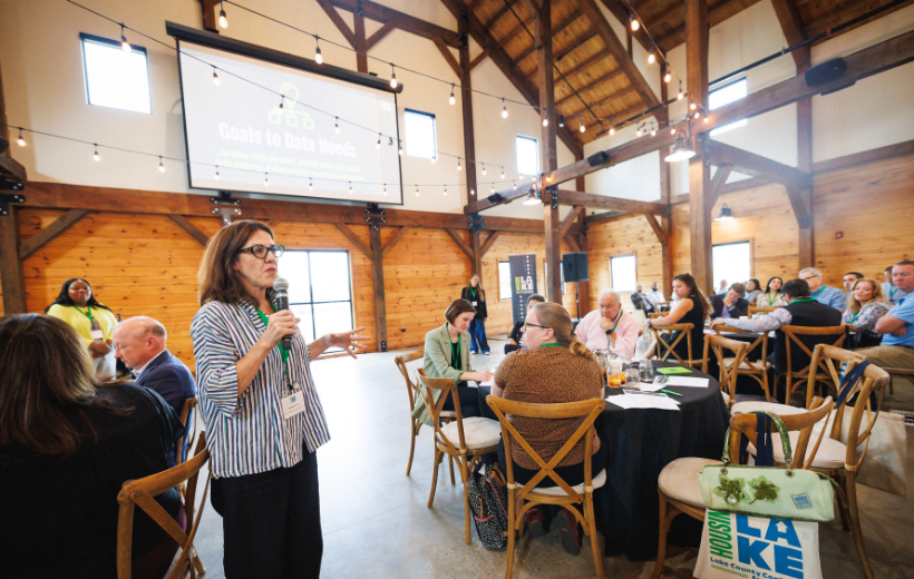 Someone holds a microphone and speaks in a room where people are seated at round tables.