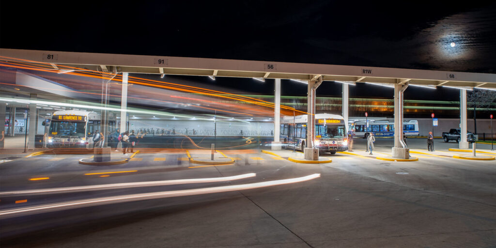 A nighttime scene at a bus and train station with buses and commuters on the move